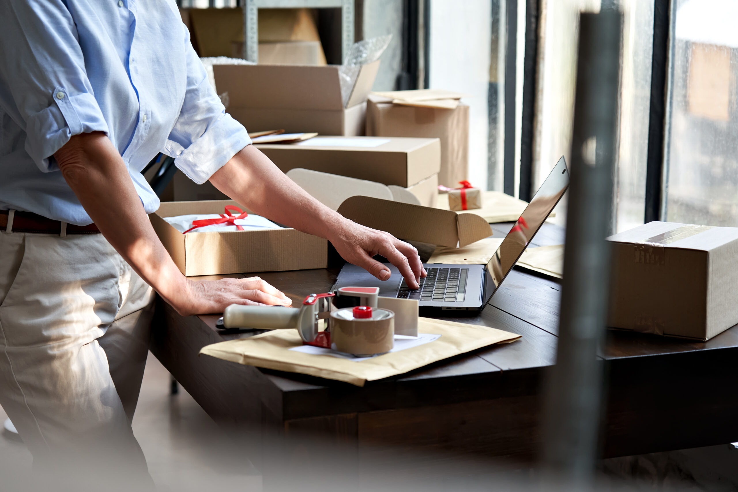 Female online store small business owner entrepreneur seller packing shipping ecommerce box checking website retail order using laptop preparing delivery parcel on table. Dropshipping concept. Closeup
